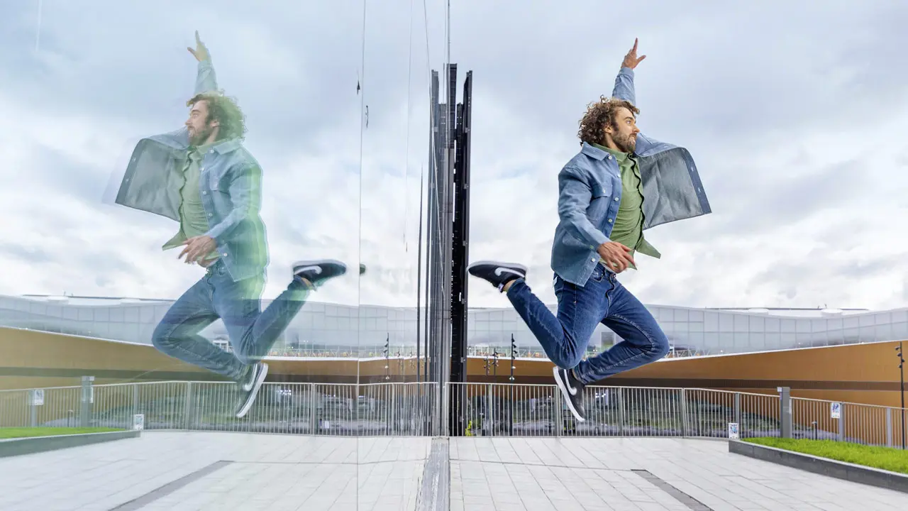 Ein Mann springt dynamisch vor einer spiegelnden Glaswand, die seine Reflexion zeigt. Er trägt eine Jeansjacke und ein grünes T-Shirt und ist in einer urbanen Umgebung mit einem modernen Gebäude im Hintergrund zu sehen.