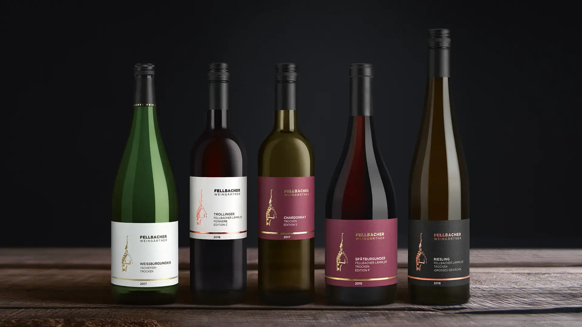 Fünf Flaschen Wein der Fellbacher Weingärtner, darunter Weissburgunder, Trollinger, Chardonnay, Spätburgunder und Riesling, präsentiert auf einem Holztisch vor einem dunklen Hintergrund.
